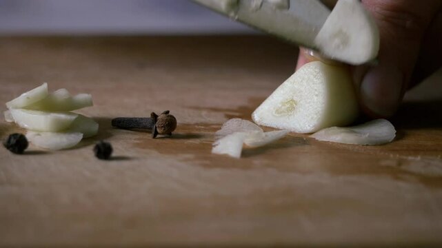 This video shows a close-up of a hand using a knife to chop garlic cloves on a wooden surface. A few black peppercorns are also visible on the surface