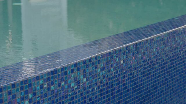 Close view of pool wall with blue mosaic tiles and flowing water. Small square tiles shine in daylight. Texture is rich with a mix of light and shadow