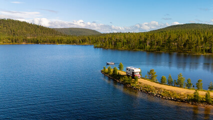 A cozy camper sits on a lush green peninsula facing a serene blue lake surrounded by dense forest....