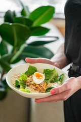 A person holding a bowl of fresh salad with lettuce, corn, tuna, and a halved soft-boiled egg. The background features green leaves, adding a natural and healthy vibe.