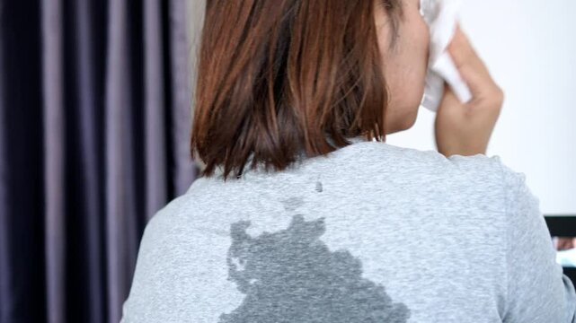 back view of asian woman sweating heavily in hot summer weather, clothes soaked from the heat, using tissue to wipe sweat from neck and face, possible signs of menopause 