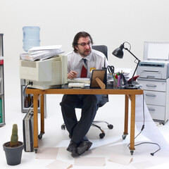 Confused office worker, man sitting in formal wear at messy desk filled with documents, gadgets and...