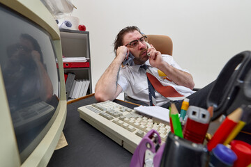 Stressed man, office worker talks on phone while holding towel to face, sitting at cluttered desk, showing chaotic multitasking and workplace discomfort. Concept of business, retro office, emotions