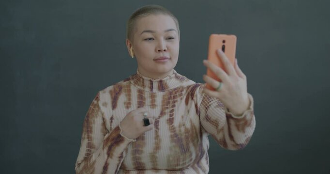 Cheerful Asian lady making online video call with smartphone and earphones talking gesturing on black color background. People and communication concept.