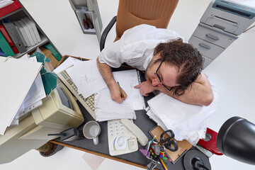 Top view of man, overworked office worker sitting at cluttered office desk, attempting to write....