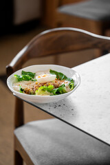 A fresh salad with lettuce, a halved boiled egg, creamy dressing, and sprinkled seasoning, served in a white bowl on a marble table, with a wooden chair in the background.
