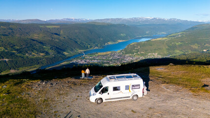 Enthusiastic travelers enjoy the stunning panorama of a Norwegian fjord, standing next to their camper van. The clear sky and green hills create a perfect backdrop for outdoor adventure.