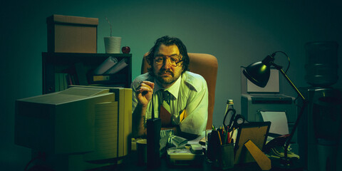 Office worker with glasses sits at cluttered desk in dimly lit room with green tint, looking...