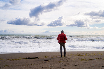 Full body back view of unrecognizable male in warm casual clothes walking alone on sandy beach towards waving stormy sea in early spring. Seascape with horizon line, sky with white clouds and human
