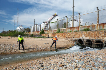 Two engineers assess water discharge from industrial drainage pipes near spherical storage tanks,...