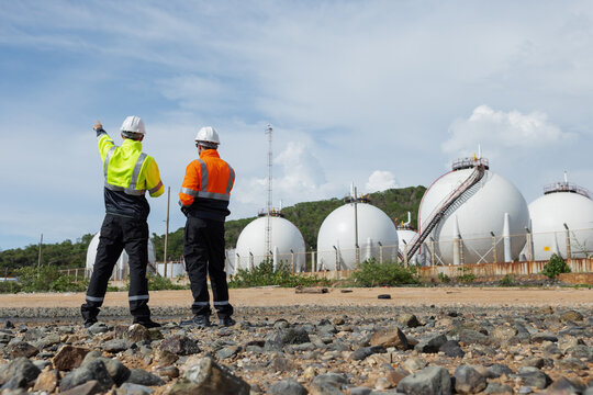 Two environmental engineers wearing safety gear observe and discuss an industrial facility with large spherical storage tanks, likely evaluating environmental safety or maintenance conditions.
