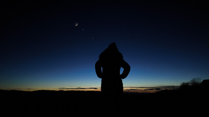 Silhouette of a woman observing Moon, planets and Milky Way stars from a dark countryside location.