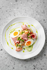 A gourmet dish featuring sliced ham, boiled eggs with runny yolks, peas, and a colorful mixed vegetable salad, elegantly plated with green sauce on a white plate against a speckled grey background