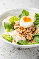 A fresh salad with romaine lettuce, soft-boiled egg, tuna, chickpeas, creamy dressing, and crispy bits, served in a white bowl on a speckled gray surface. Healthy and delicious meal.