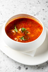 A bowl of hot, spicy soup garnished with fresh cilantro and green herbs, served on a white plate against a speckled gray background. 
