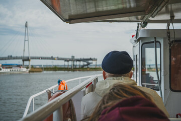 An elderly man in a black cap sits on a bench on a ship that sails on the Masurian lakes. View from behind.