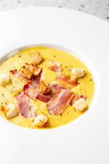 A bowl of creamy yellow soup with crispy bacon and croutons, elegantly served on a white plate against a textured gray background. Gourmet presentation, top view.