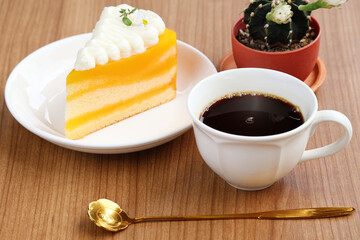 Hot black coffee with slight foam in white cup placed between orange cake with whipped cream and small potted cactus with white flowers on wooden table.

