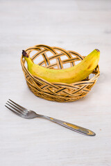 Fototapeta premium A single, ripe yellow banana rests in a small, woven basket on a white wooden table. A metal fork is placed next to the basket, suggesting a simple meal or snack.