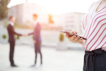 Woman is holding a mobile phone in the background handshake of two business men. Blurred business people on the background. Overhear.
