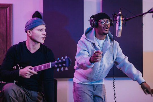 Two musicians passionately perform in a recording studio, one playing guitar and the other singing into a microphone. The vibrant atmosphere highlights creativity and teamwork in music production.