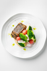A gourmet dish featuring sesame-crusted fish, fresh broccoli, radish slices, and cherry tomatoes, elegantly plated with sauce and green oil droplets on a white plate.