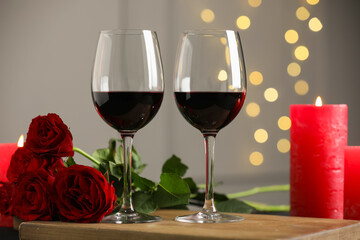 Glasses of red wine, burning candle and roses on table against gray background with blurred lights. Romantic dinner