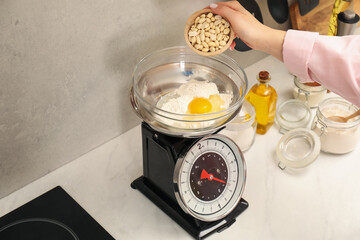 Woman adding peanuts into bowl on mechanical kitchen scale at white marble countertop indoors, closeup