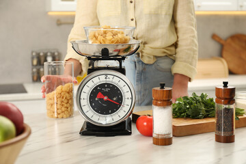 Woman holding container of raw pasta near mechanical kitchen scale at white marble countertop...