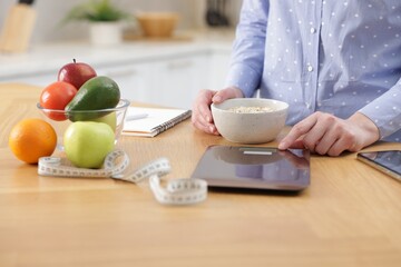 Woman weighting oatmeal on kitchen scale at wooden table indoors, closeup