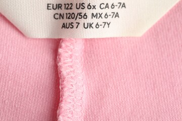 Clothing label with size on pink garment as background, closeup