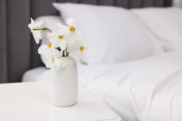 Beautiful daffodils in vase on white table near bed at home. Space for text