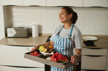 A cheerful woman in a striped apron holds a tray with fresh fruits, including watermelon,...
