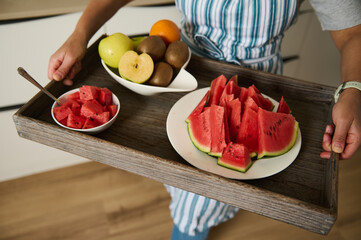 A close-up of a person holding a wooden tray with fresh fruit, including vibrant watermelon slices...