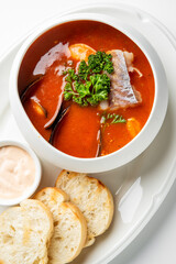 A bowl of seafood soup with mussels, shrimp, and fish in a tomato-based broth, garnished with parsley. Served with toasted bread and a creamy dipping sauce on a white plate.