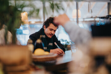 A man is seated at a table in a cozy cafe, writing attentively. Warm lighting and a relaxed...