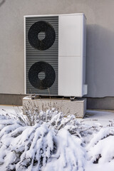 Close-up of a dual-fan outdoor heat pump on a gray wall with snow-covered bushes, highlighting...
