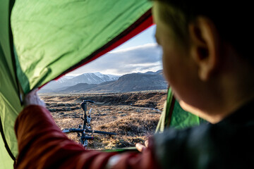 A view of mountain range in Iceland from inside a tent, a woman is holding the tent door open and a bike is lying on the ground in front of the tent.
