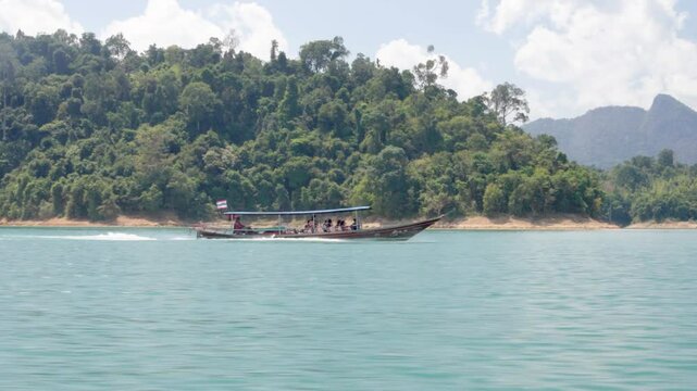 A traditional Thai Longtail boat with tourists sails through the bays of Cheo Lan Lake in Kaosok Park against the background of green islands, side view