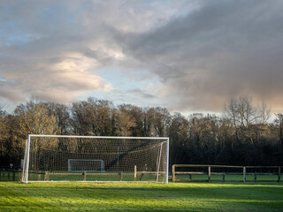 Fototapeta premium A soccer field with a goal and a fence in the background. The sky is cloudy and the grass is green. Football training ground. Popular outdoor sport. Nobody.