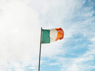 Irish national flag waving on a pole, blue cloudy sky background.