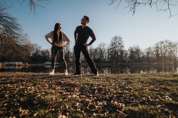 A couple stands together in a park during sunset, enjoying the serene scenery and shared moment by a peaceful lake with trees in the distance.