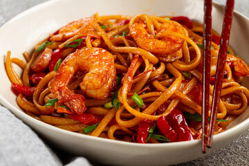 udon stir-fry shrimp noodles with red bell pepper and green onion, served in a black bowl, with chopsticks, gray background,