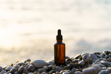 Brown glass jar of serum on pebbles on sea water background with sunset light. Concept of beauty products, skin care, hydration