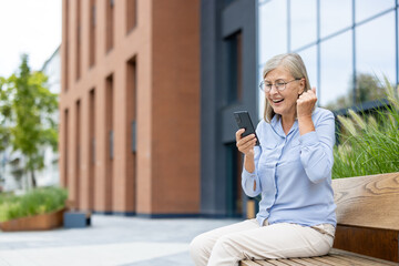 An excited senior woman wearing glasses looks at her phone, celebrating good news while sitting...