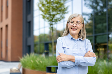 A smiling senior woman with crossed arms poses outdoors near a modern building with glass windows.