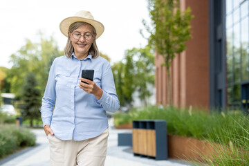 A smiling senior woman in a hat uses her phone while walking outdoors in a modern urban setting,...