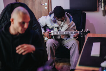 Two musicians in a recording studio work together. One plays the electric guitar while the other listens intently. The setting is modern and creative, fostering an atmosphere of musical collaboration.