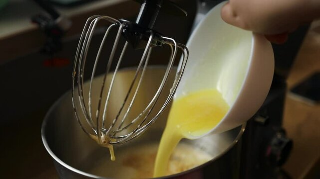 Close-up view of liquid mixture, possibly butter and eggs, being poured into a stand mixer bowl with whisk attachment. A key moment in dessert or bread baking, perfect for recipes and food tutorials