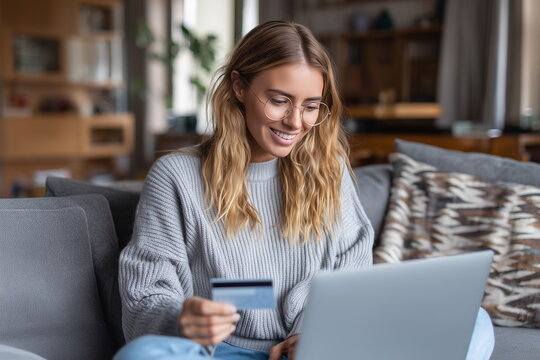 A young White woman sitting on the sofa at home in a living room with a laptop on the lap and holding a credit card. Online shopping, payment, purchase, buy. E-commerce. - Powered by Adobe